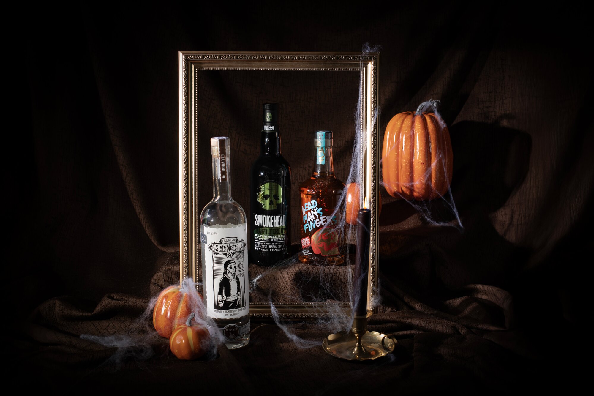 Three bottes of spirits on a dark background surrounded by pumpkins and spiderwebs. 
