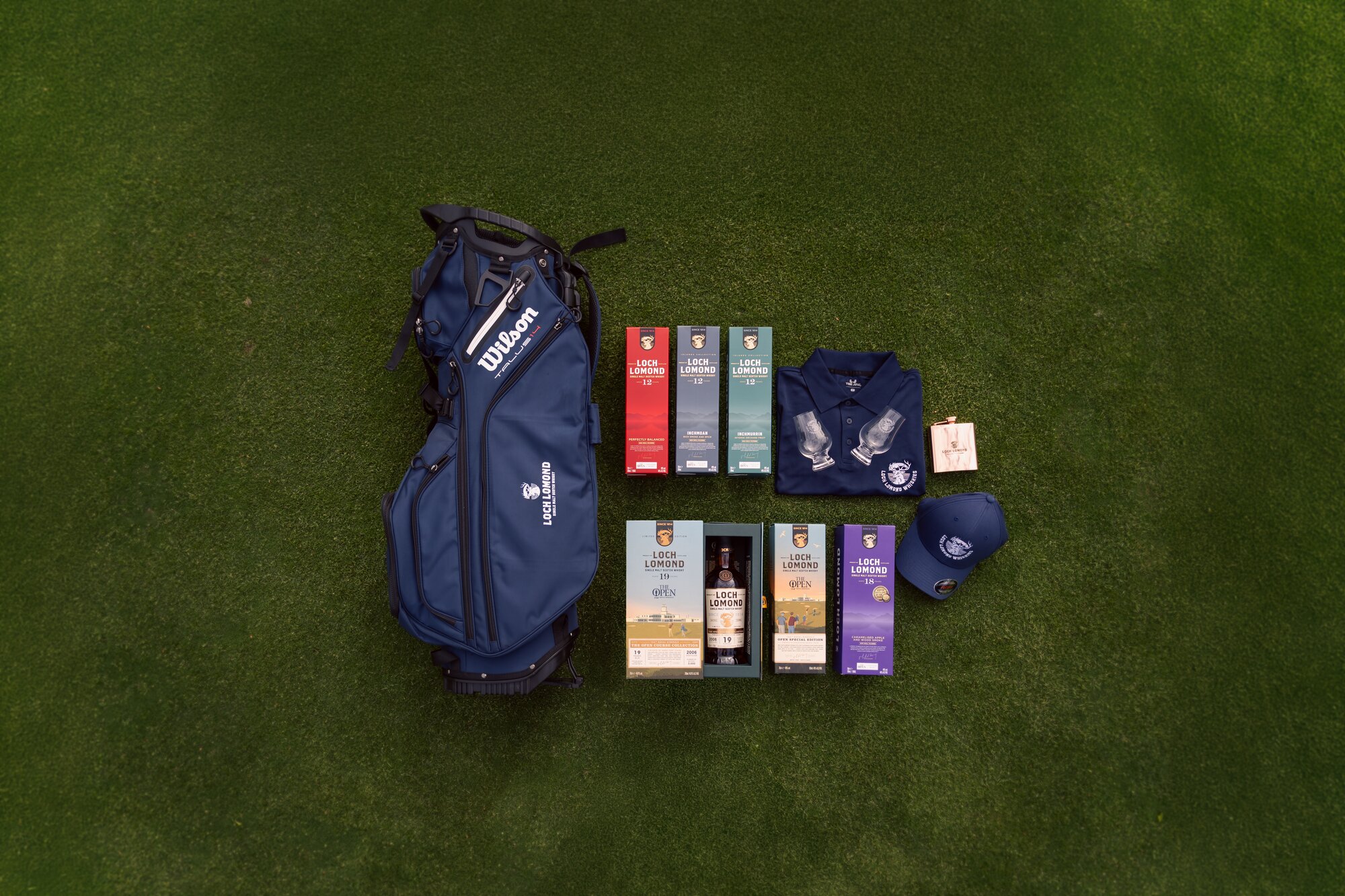 A golf bag with bottles of whisky, a hat, flak, polo t-shirt, and glasses laid out on grass. 
