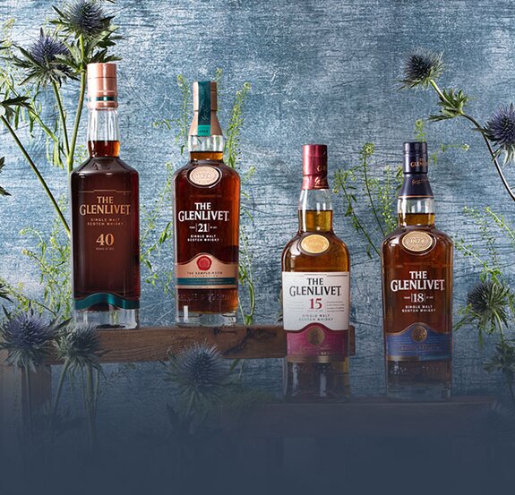 Four bottles of Glenlivet whisky surrounded by thistles on a blue background.