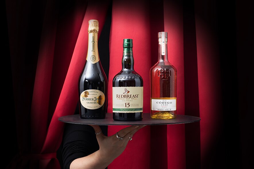 A bottle of Perrier-Joet, Redbreast 15 and Codigo Rosa on a black tray in front of red stage curtains. 