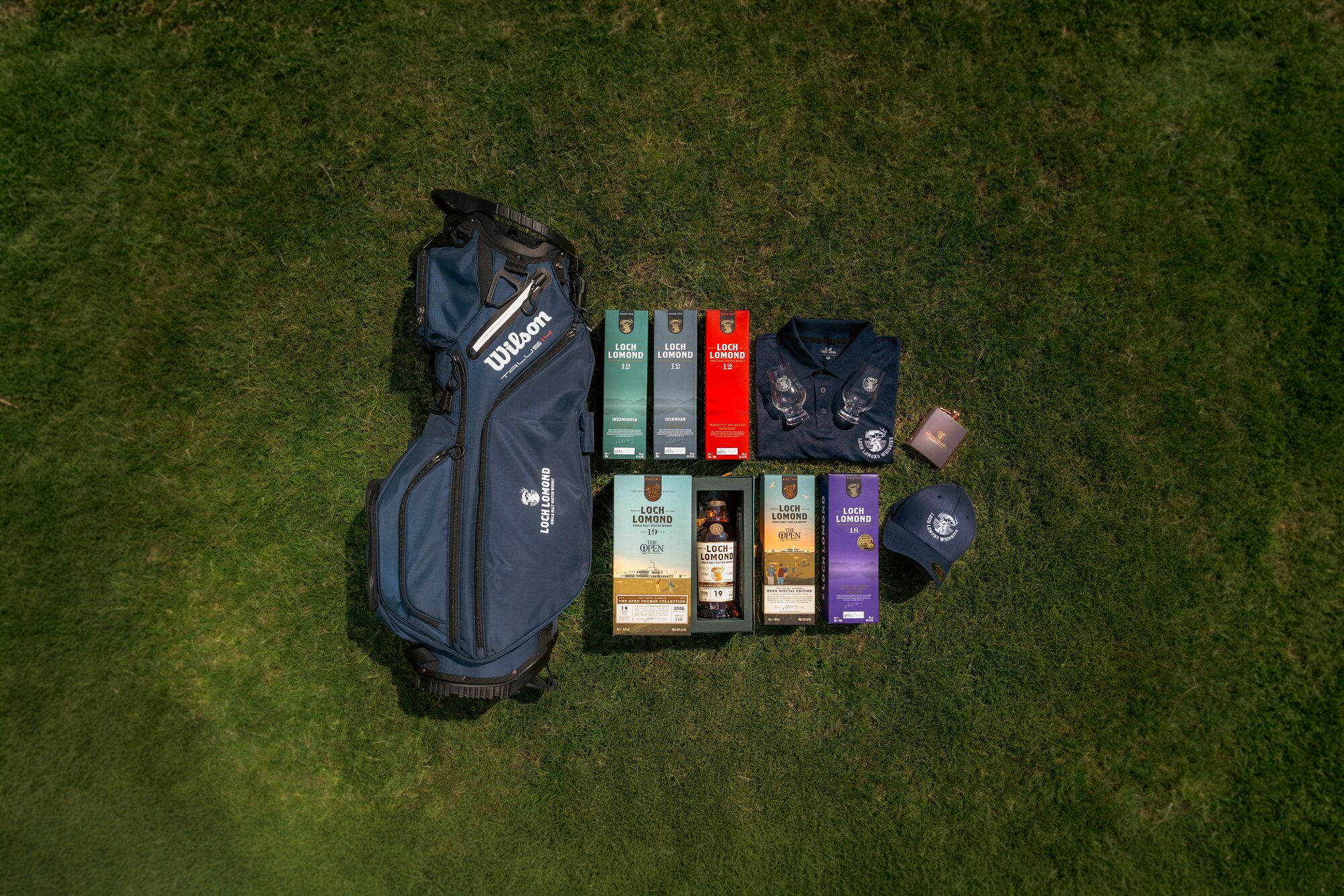 A golf bag with bottles of whisky, a hat, flak, polo t-shirt, and glasses laid out on grass. 