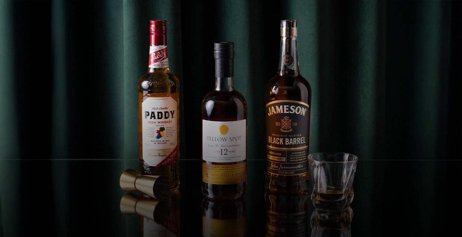 Bottles of Paddy's Irish Whiskey, Yellow Spot 12 Year Old and Jameson Black Barrel on a reflective surface with a green curtain backdrop.
