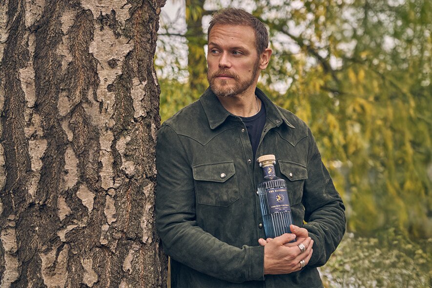 Sam Heughan holding a bottle of Sassenach vodka leaning against a tree.