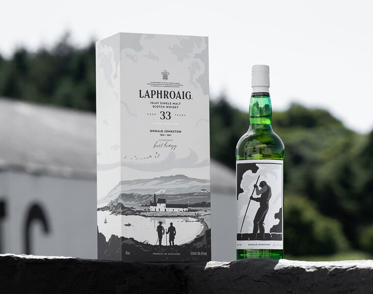 A bottle of Laphroaig 33 Year Old Strong Characters next to its presentation box with the distillery in the background. 