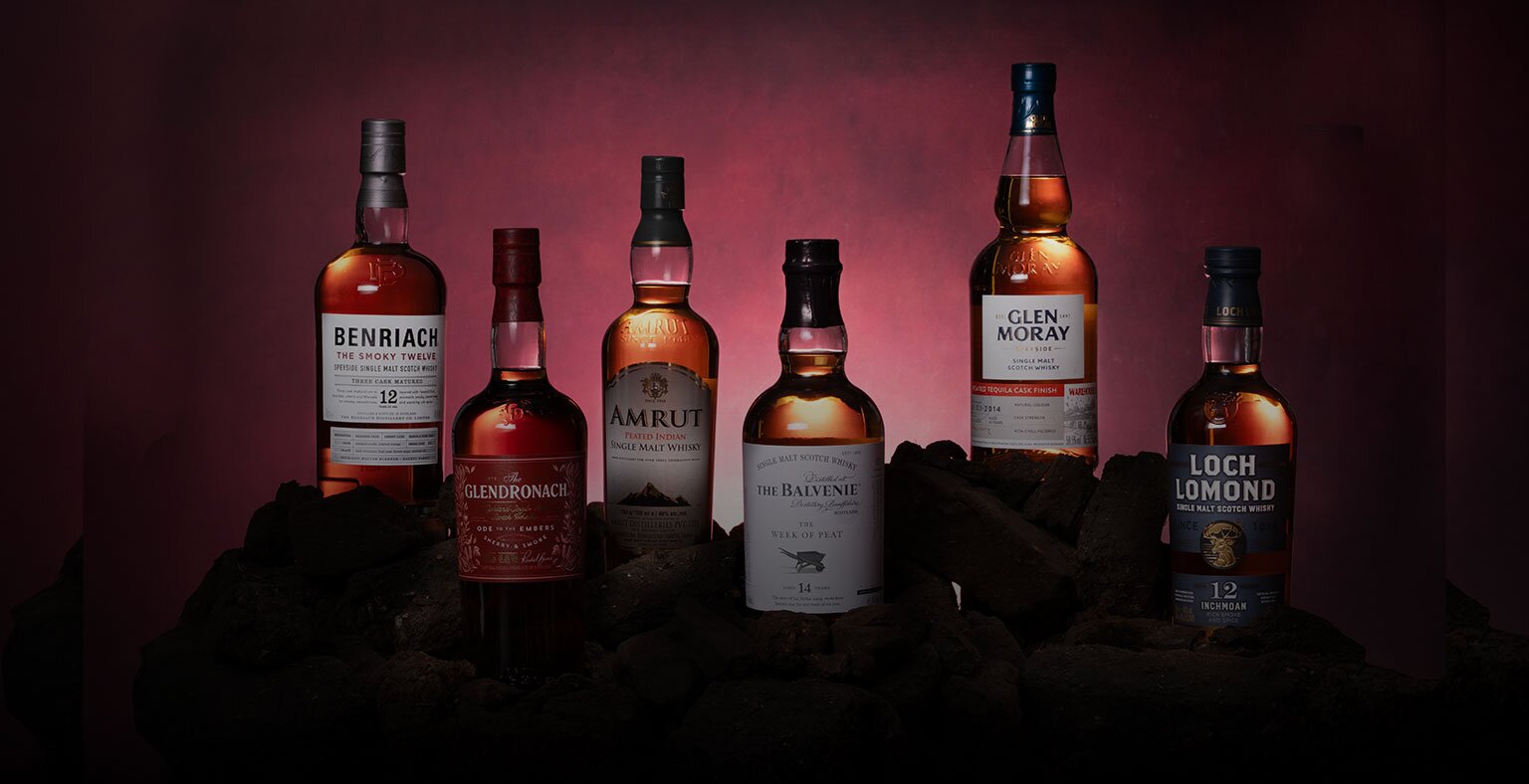 Six bottles of peated whisky against a red background surrounded by peat bricks
