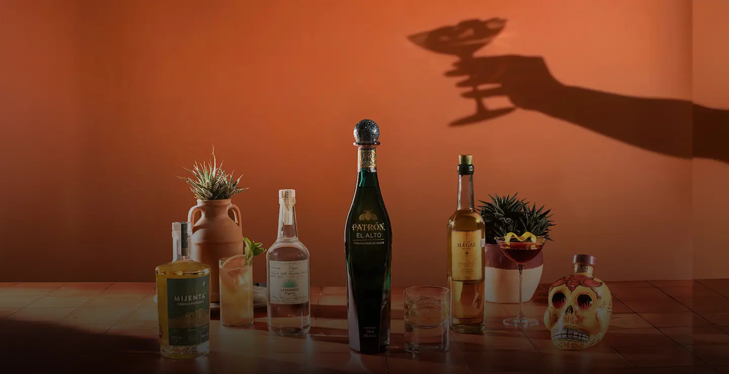 Five bottles of Tequila and Mezcal against a dusky orange background with a shadow holding a cocktail.