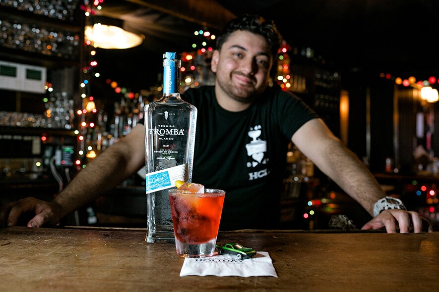 A bottle of Tromba Tequila Blanco on a bar next to a cocktail.