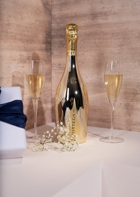 A bottle of Bottega Gold Prosecco and two champagne flutes against a pink and beige background. 