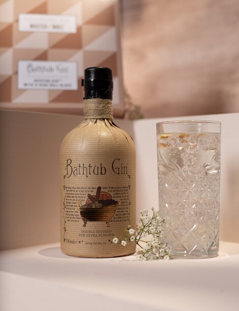 A bottle of Bathtub Gin, highball glass and gift set box against a pink and beige background. 