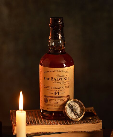 A bottle of Balvenie 14 Year Old Caribbean Cask whisky on top of a stack of books next to a candle. 