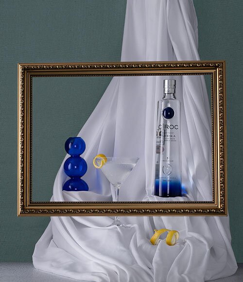 A bottle of Ciroc next to a martini cocktail behind a gold picture frame.