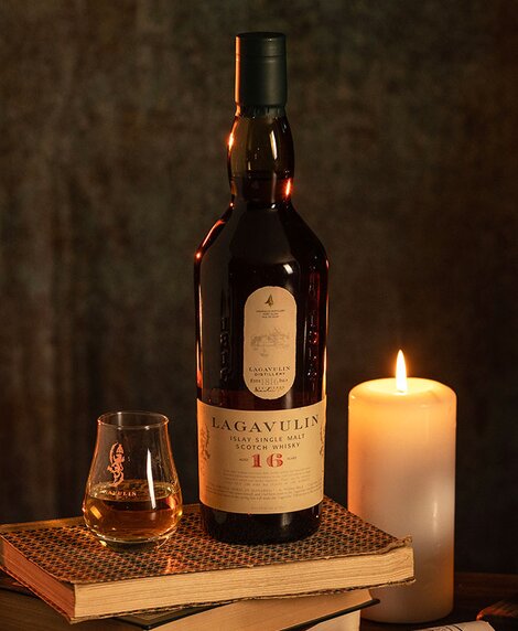 A bottle of Lagavulin 16 year old whisky on top of a stack of books next to a candle. 
