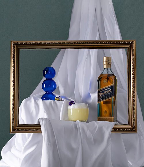 A bottle of Johnnie Walker Blue Label next to a Whisky Sour behind a gold picture frame.