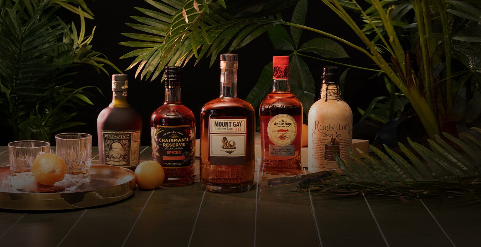 Five bottles of rum with tropical plants in the background, and a glass of rum to the left.