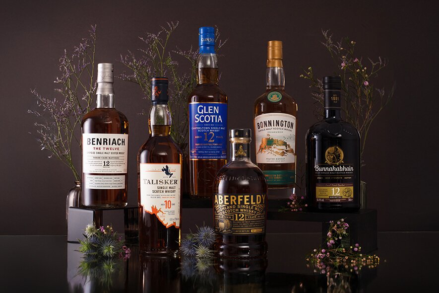 Six bottles of Scotch whisky on a dark background surrounded by delicate flowers.