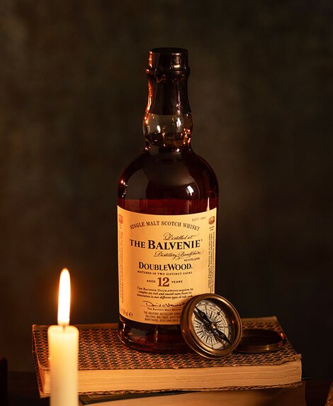 A bottle of Balvenie Doublewood 12 Year Old whisky on top of a stack of books next to a candle. 
