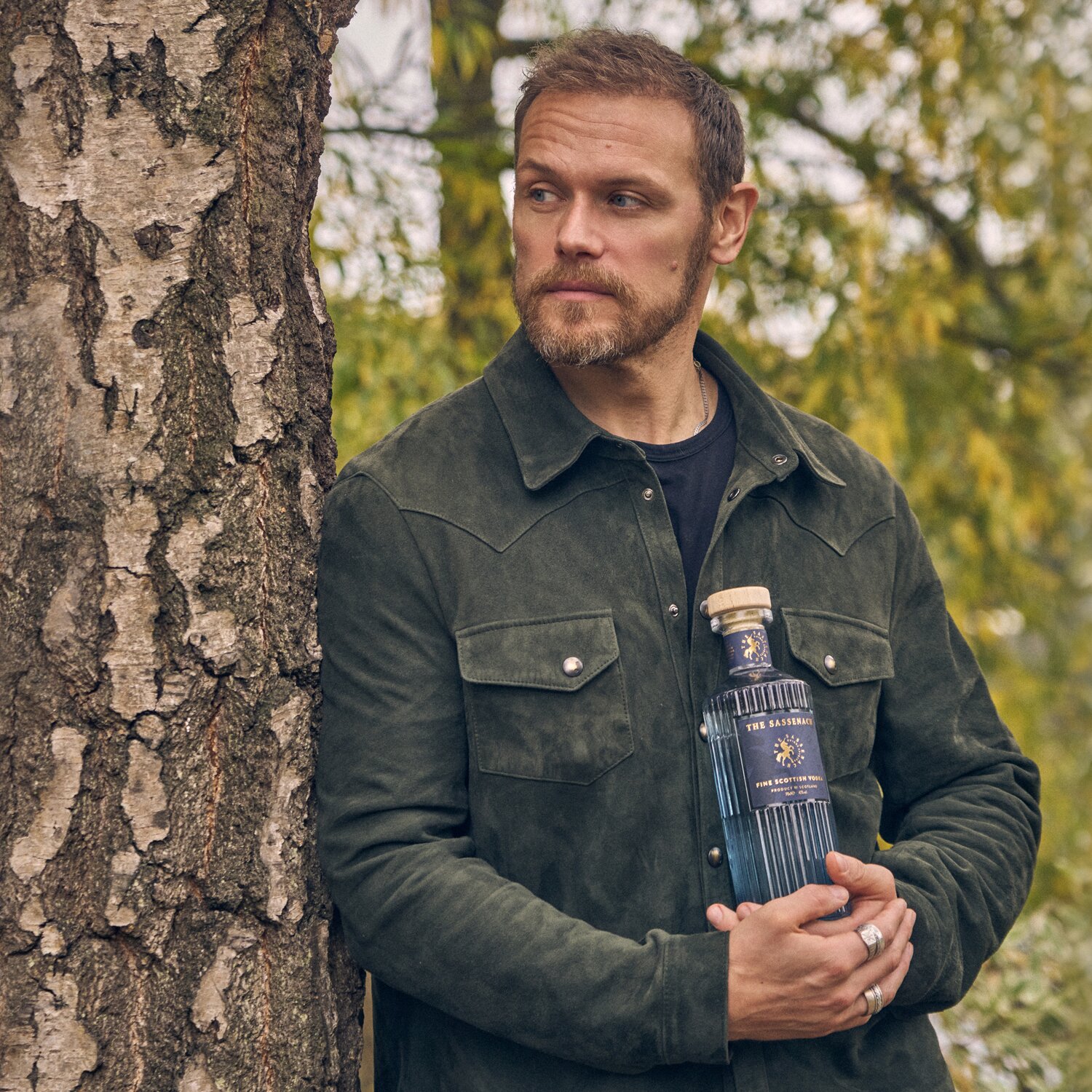 Sam Heughan holding a bottle of Sassenach Vodka next t a tree.