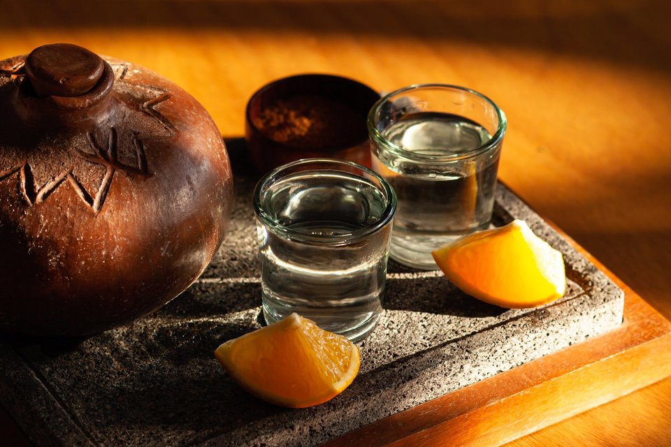 Two shot glasses full of blanco Tequila next to some traditional pottery, with two wedges of orange alongside.