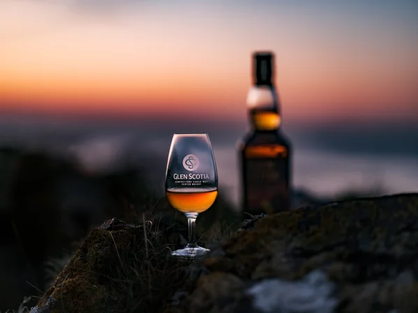 A tasting glass of whisky on a beach with a bottle of Glen Scotia 14yo Icons #2 whisky out of focus in the background.