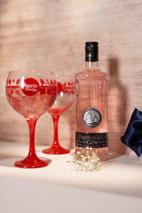 A bottle of Puerto De Indias Strawberry Gin and two gin bowls against a pink and beige background. 