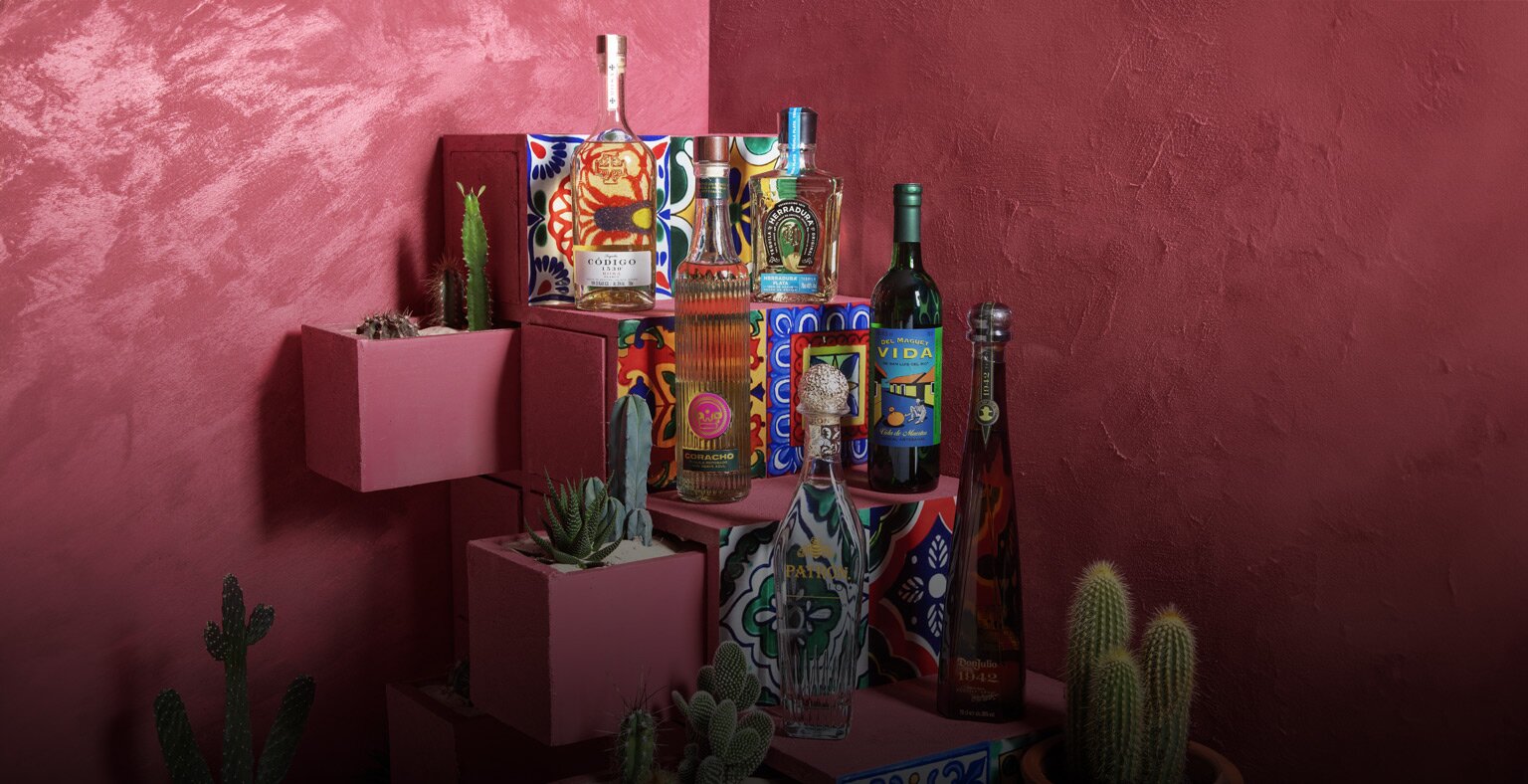 Sixe bottles of tequila and mezcal on a pink stair case surrounded by colourful Mexican tiles and cacti. 