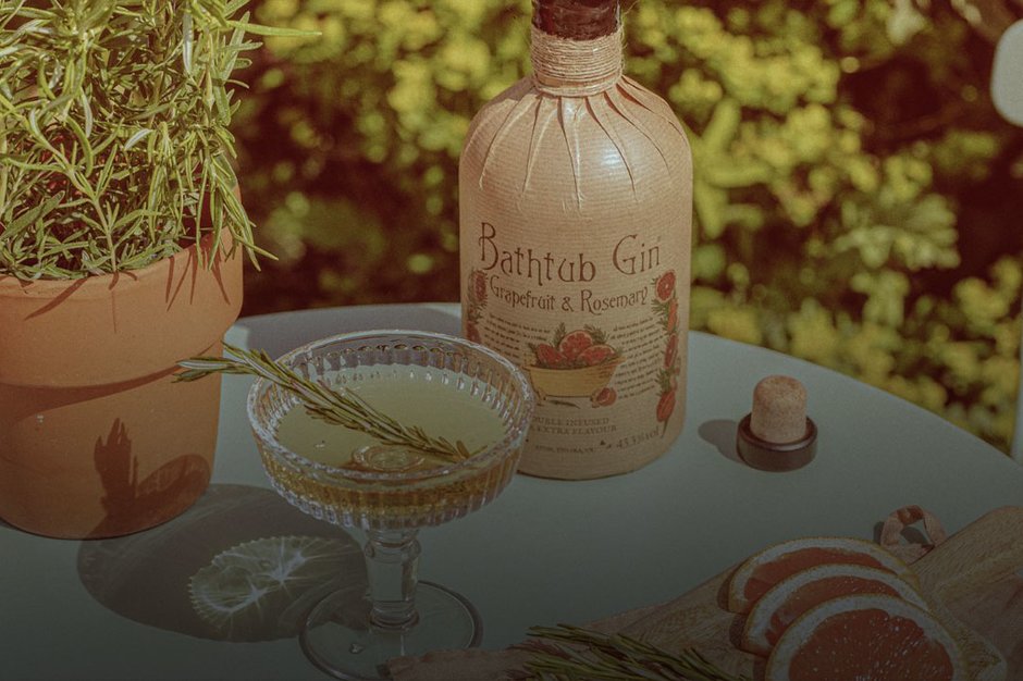 A bottle of Bathtub gin on a table with a cocktail, grapefruit slices and a Rosemary sprig.