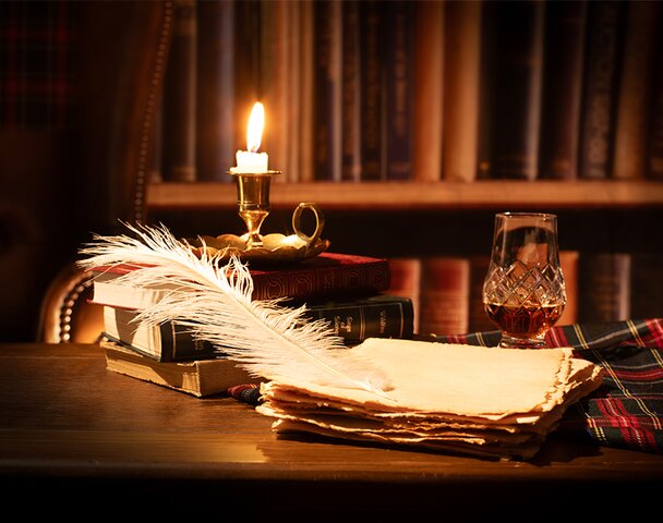 A desk with a a quill, stack of paper and glass of whisky on it.