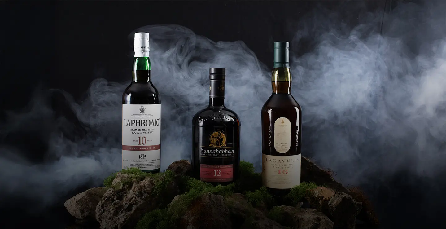 Three bottles of Islay whisky on a mossy rock surface surrounded by fog.