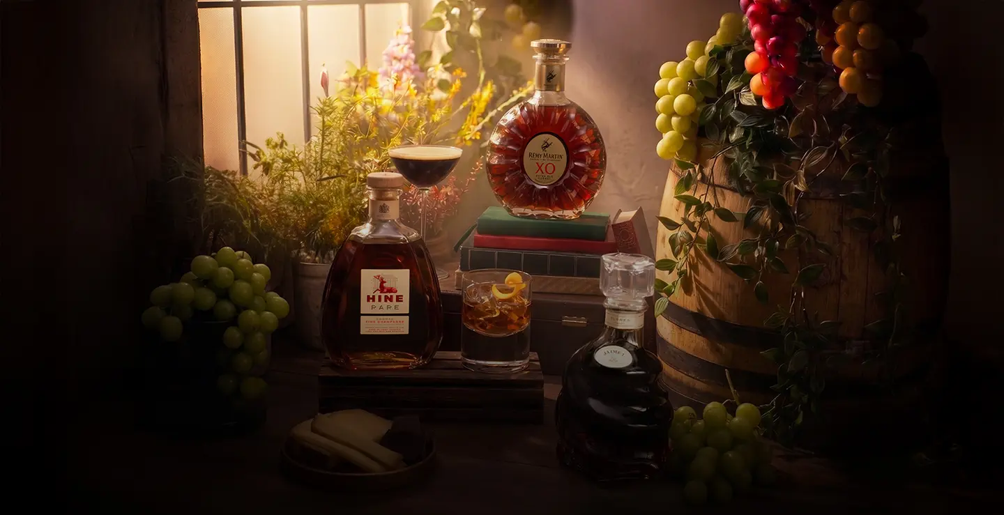 Three bottles of brandy next to a sunny window surroudned by greenery and grapes.