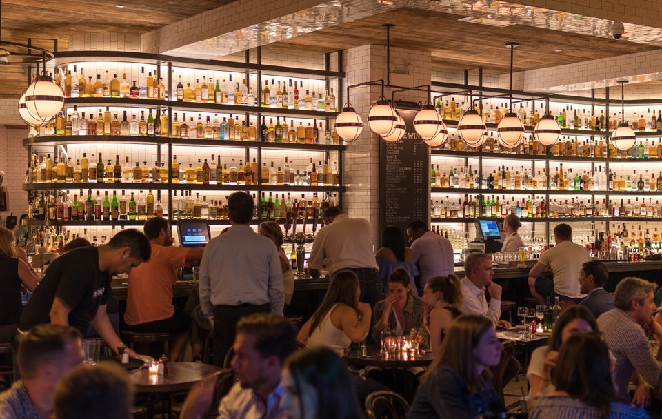 A busy, warmly lit bar with industrial and art deco features and featuring large wall filled with bottles of alcohol.