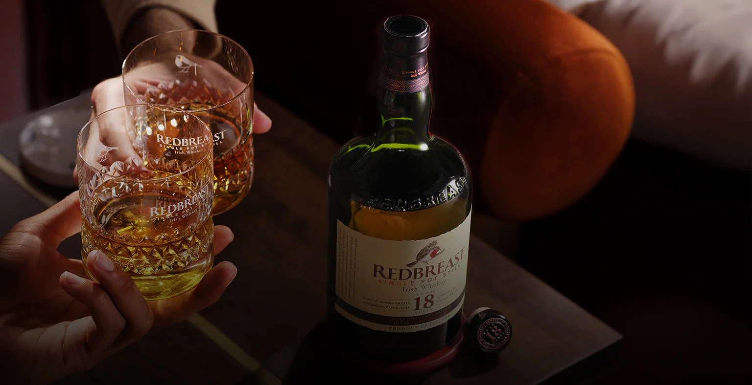 A bottle of Redbreast 18 year old whisky next to two people holding glasses of whiskey.