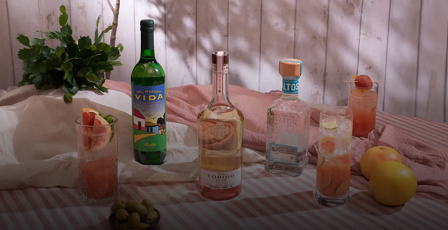 A bottle of Codigo Rosa, Del Maguey Mezcal and Altos tequila on pink stiped tablecloth next to grapefruits and Paloma cocktails.