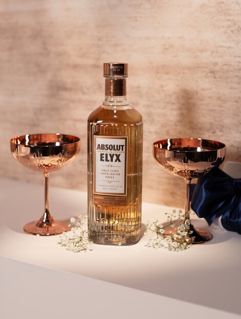 A bottle of Absolut Elyx vodka and two metal coupe glasses against a pink and beige background. 