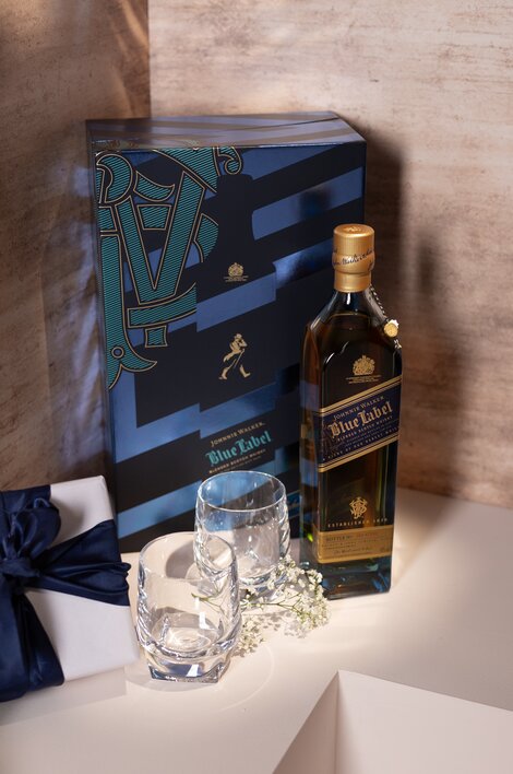 A Johnnie Walker Blue Label bottle, gift set box and pair of crystal glasses against a pink and beige background. 