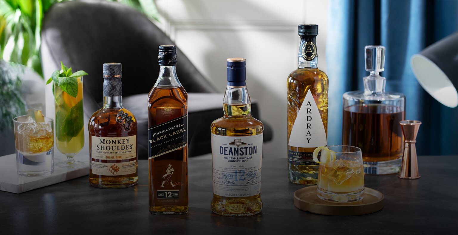 Four bottles of whiksy next to cocktails on a table with cream background.