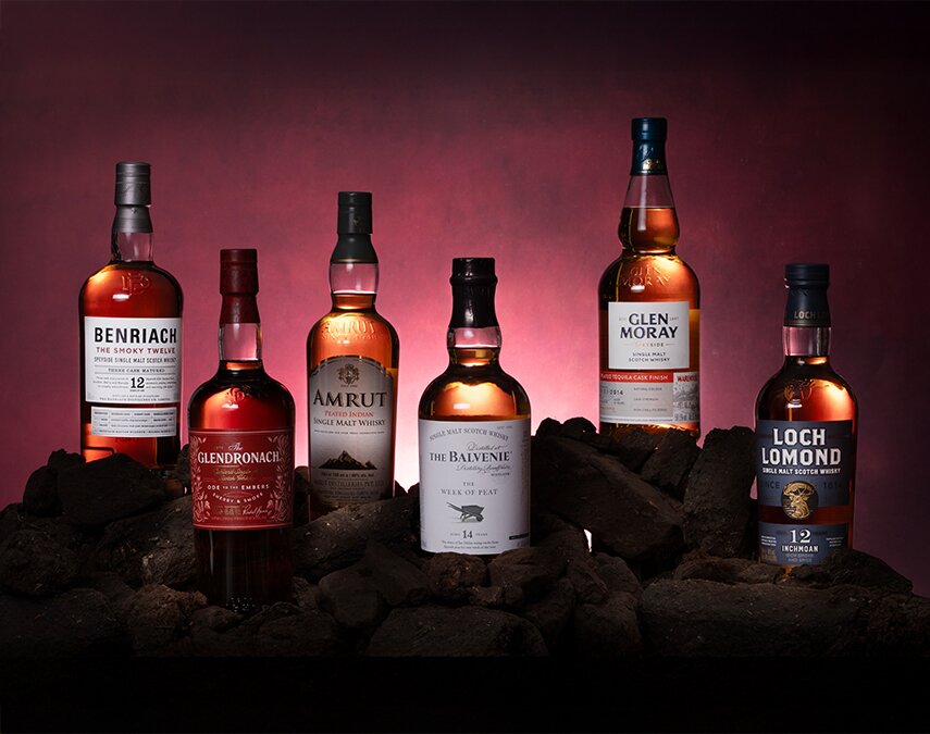 Six bottles of peated whisky against a red background surrounded by peat bricks