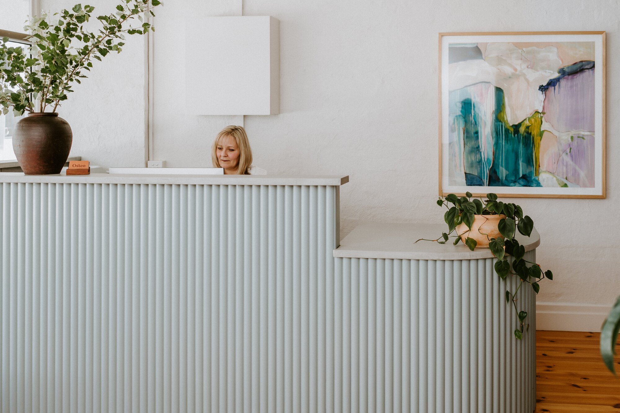 A photo of the Rhythm Health reception desk, with our receptionist, Sharyn working behind it. The desk has several potted plants and flower arrangements on it.