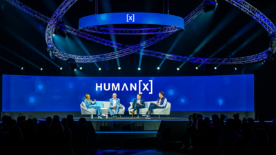 A panel of four speakers is seated on a stage with blue lighting and a large screen behind them displaying the conference name, "HUMAN [X]." The setting includes modern seating and overhead circular truss lighting. The audience is visible in the foreground.