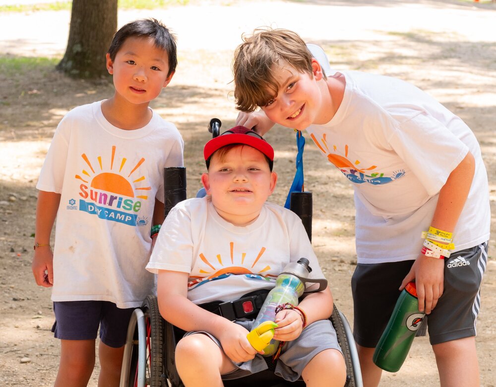 Three children, one in a wheelchair in the middle, smile at the camera at camp