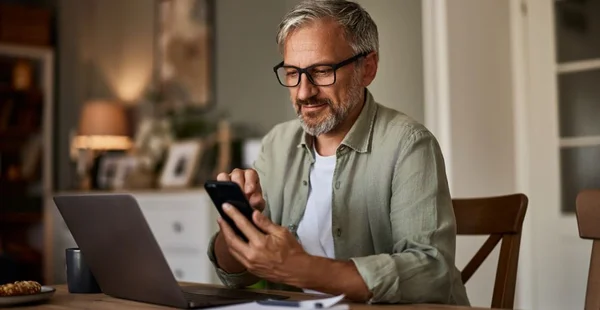 Um freelancer sorridente com óculos usando um telefone enquanto trabalha em um laptop.