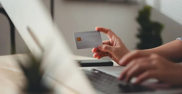 Foto em close-up das mãos femininas segurando cartão de crédito e usando laptop para pagamento online em home office.