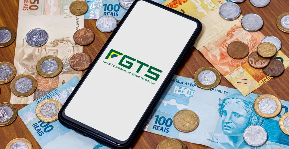 Logotipo do FGTS na tela do smartphone com diversas moedas e dinheiro ao redor. O FGTS é o fundo de garantia do trabalhador brasileiro. Pagamento pelo governo brasileiro