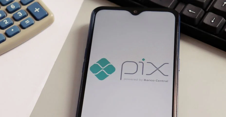 Logotipo do Pix na tela do smartphone, com computador e calculadora ao fundo. Pix Brasil é um sistema de pagamento instantâneo desenvolvido pelo Banco Central