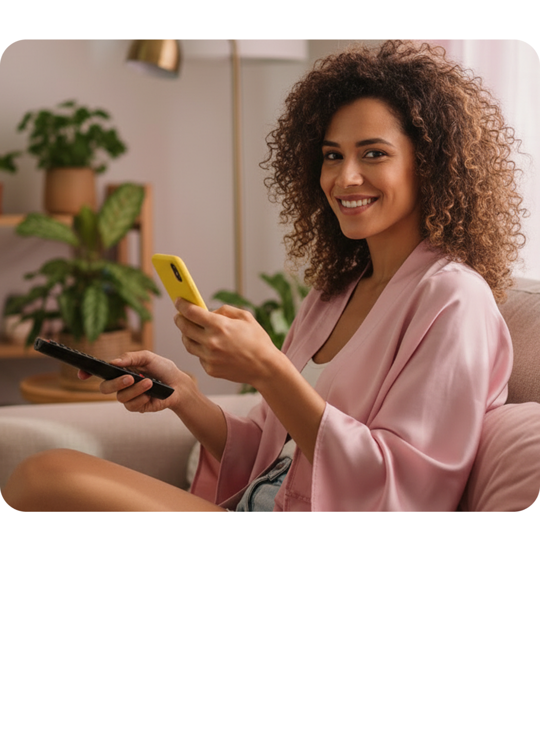Pessoa sentada em um sofá, segurando um smartphone amarelo em uma mão e um controle remoto na outra, usando um roupão rosa.