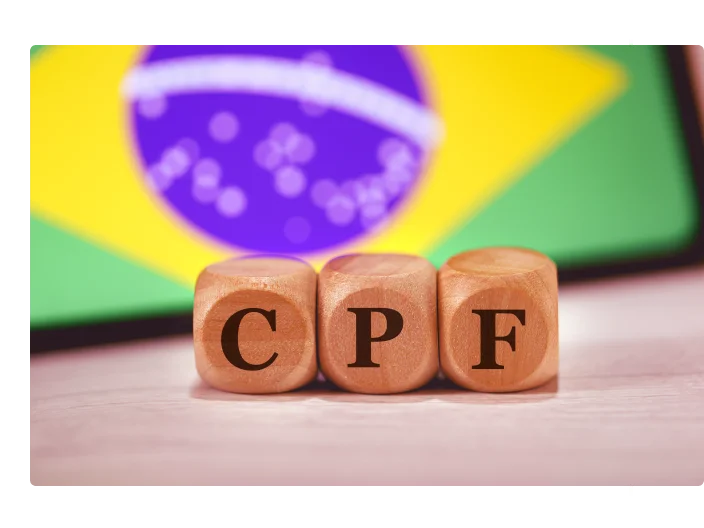 A sigla CPF para Registro de Pessoa Física em português brasileiro escrita em dados de madeira. Ao fundo, a bandeira do Brasil na tela de um celular.
