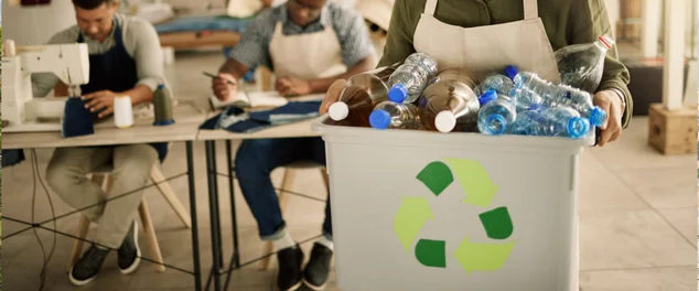 Costureira alegre segurando um balde de garrafas plásticas recicladas. Designer mestiço segurando lixo de garrafas plásticas renováveis. Jovem alfaiate reciclando garrafas plásticas de seu estúdio