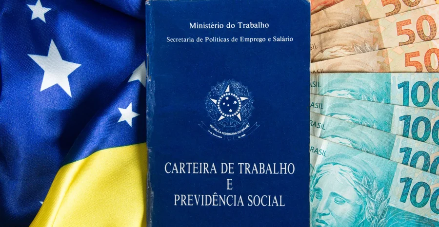 Carteira de Trabalho e Previdência Social com cédulas de dinheiro brasileiras e bandeira brasileira em segundo plano