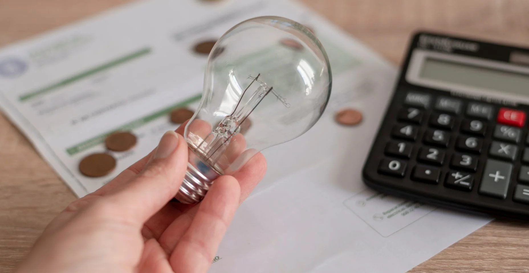 Uma mão segurando uma lâmpada sobre a conta de luz, várias moedas e uma calculadora na conta. Conceito de preços de eletricidade e pagamento de impostos.
