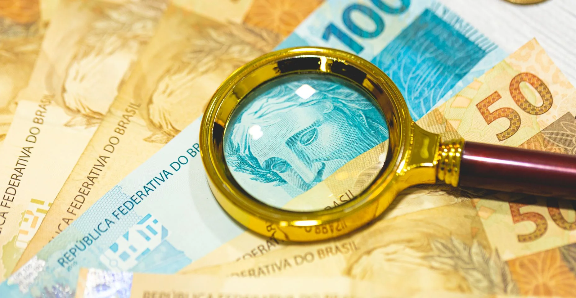Notas de reais sobre uma mesa de madeira com uma lupa dourada na composição. Economia brasileira, finanças e inflação