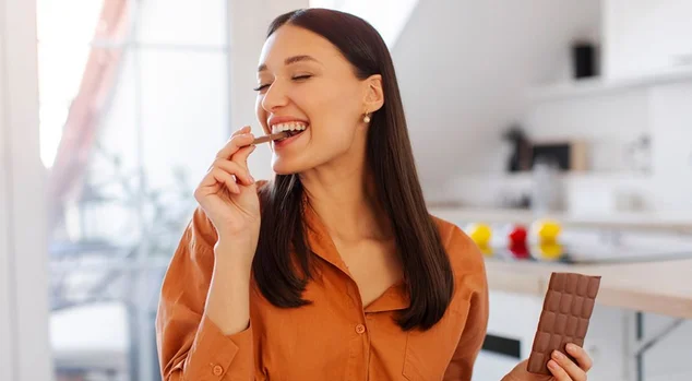 Mulher feliz com olhos fechados desfrutando do sabor da barra de chocolate, sentada na cozinha em casa. Guloso, amor por doces e doces